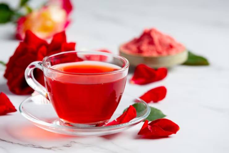 Glass cup of aromatic rose tea with vibrant red infusion, surrounded by fresh rose petals and dried rose powder, highlighting herbal tea benefits and natural wellness.
