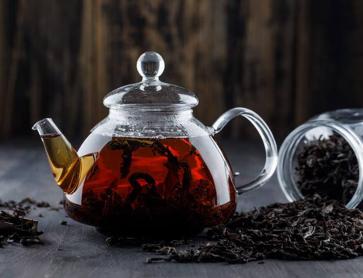 Glass teapot filled with freshly brewed black tea, surrounded by loose black tea leaves on a dark surface.