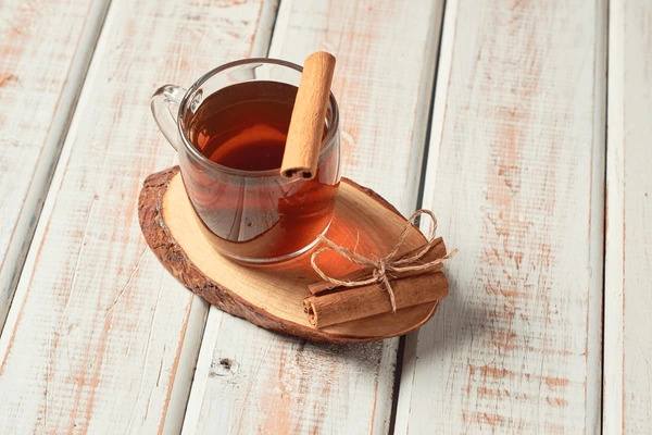 Glass cup of cinnamon tea with a cinnamon stick, served on a wooden coaster over a rustic wooden background.