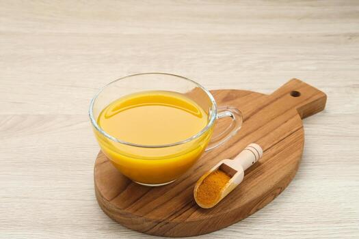 Warm turmeric tea in a glass cup on a wooden board with turmeric powder scoop on a light background.