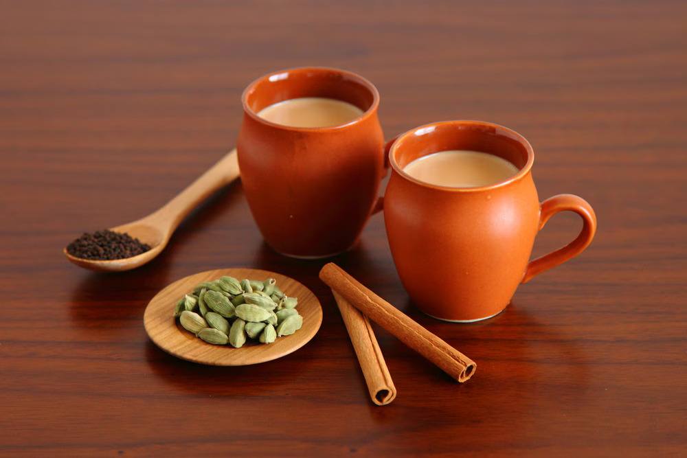 Two cups of Indian elaichi chai tea served with cardamom pods, cinnamon sticks, and black tea leaves on a wooden table